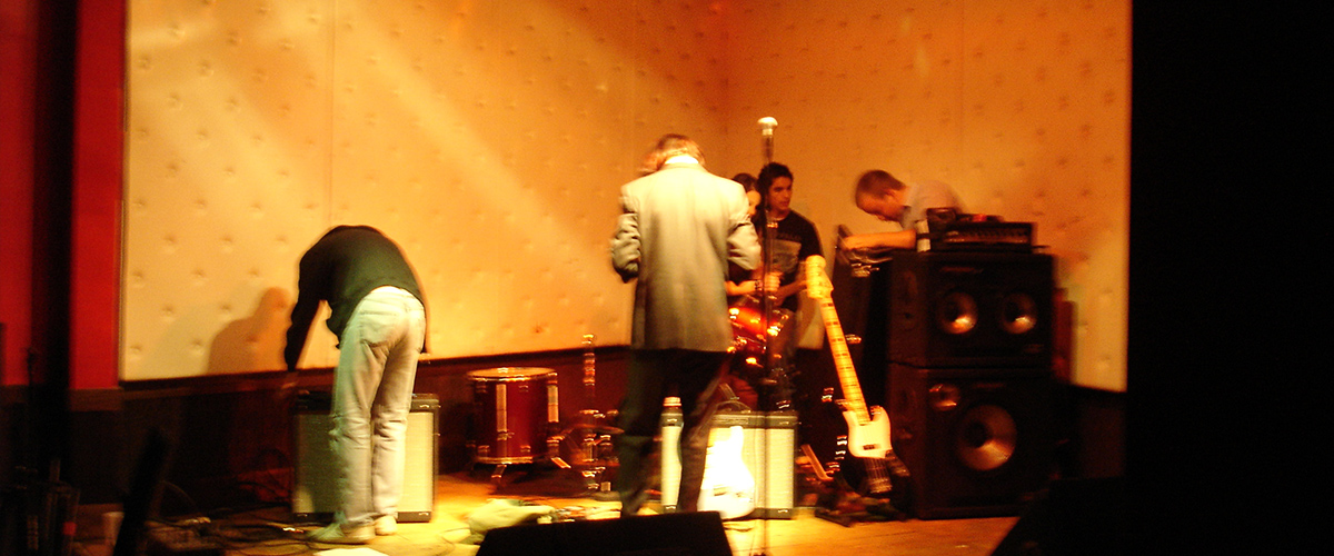 About Header Image. The band setting up their equipment on stage.